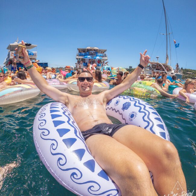 A man wearing sunglasses and swim trunks relaxes on an inflatable pool float in the water, surrounded by other people on floats and boats, under a clear blue sky at a lively yacht party.