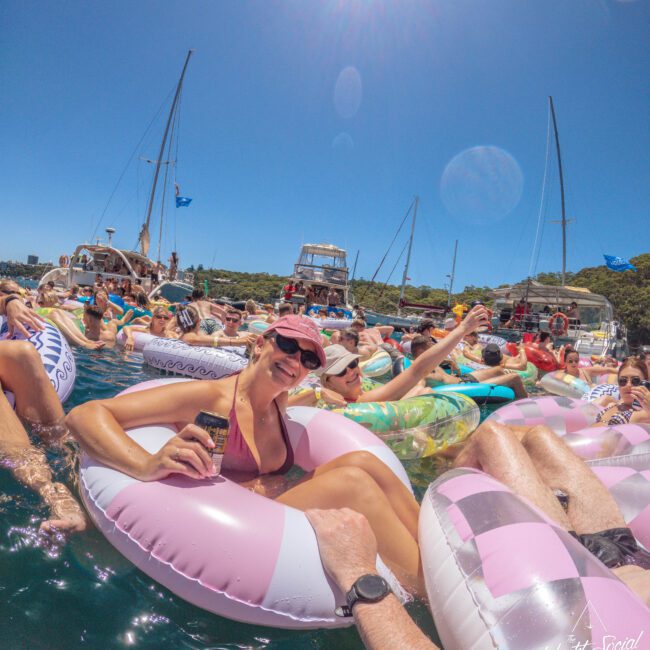 A group of people relax on inflatable pool floats in the water, holding drinks and smiling under a clear blue sky. Sailboats are anchored nearby, and the atmosphere is festive and sunny.