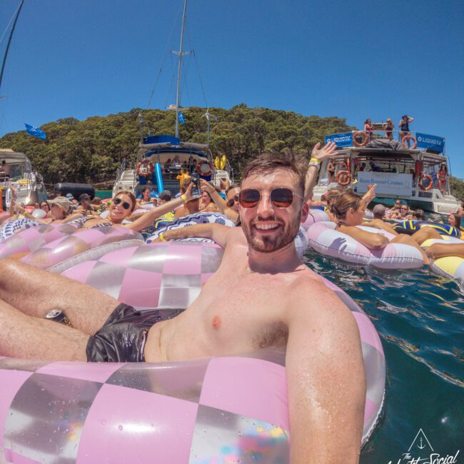 A man wearing sunglasses smiles while lounging on a pink and white inflatable float in the water, surrounded by other people on floats. Boats and green trees are visible in the background under a clear blue sky.