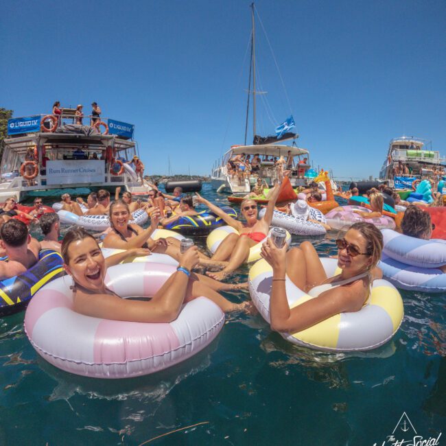 People relax and smile on colorful inflatable pool floats in the water, holding drinks, surrounded by boats and others at a lively social gathering under a sunny blue sky.