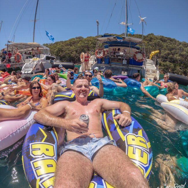 A man in swim trunks smiles and holds a can while relaxing on a yellow and blue inflatable in the water, surrounded by other people on colorful floaties near yachts on a sunny day.