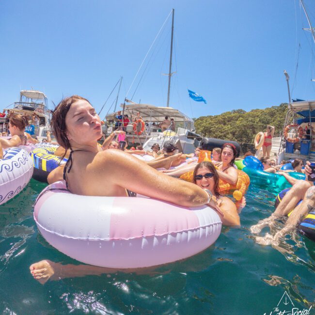 Several people relax on colorful inflatable tubes in the water near docked boats under a clear blue sky. A woman in the foreground sits in a pink float, while others chat and enjoy the sunny day.