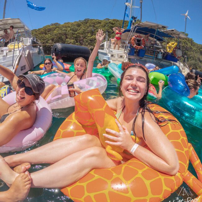 A group of smiling young adults relax on colorful inflatable pool floats in the water near boats on a sunny day. One woman in front holds a giraffe-shaped float and others wave, enjoying the lively atmosphere.