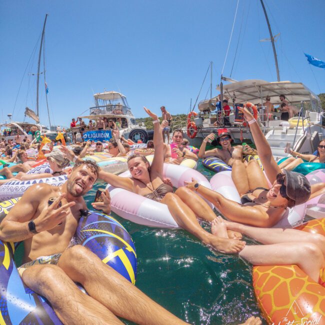 A group of people relax on colorful pool floats in the water, smiling and posing for the camera, with boats and a clear blue sky in the background.
