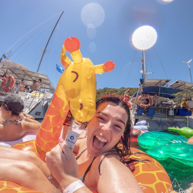 A smiling woman in a pool floats on an inflatable giraffe, holding a drink can. She is surrounded by boats and other people enjoying a sunny day on the water.