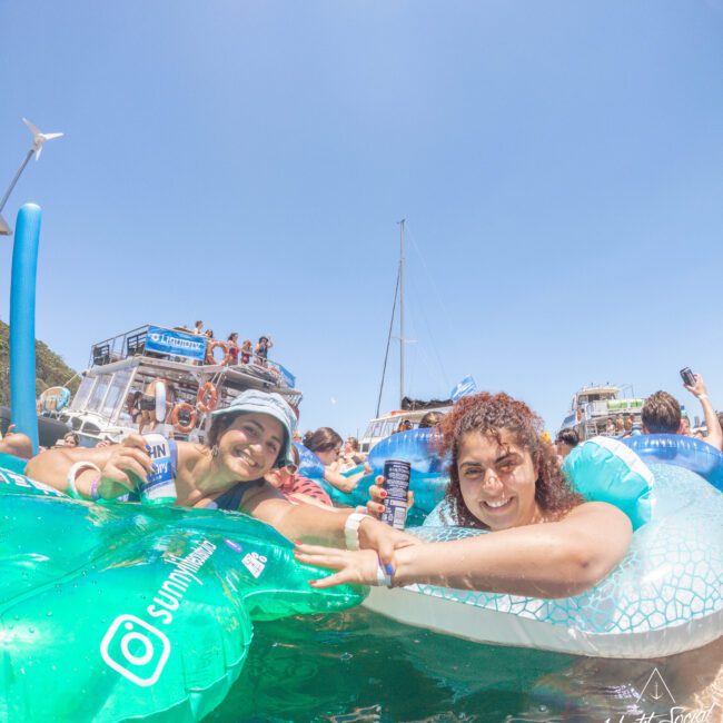 Two women smile and hold drinks while relaxing on inflatable pool floats in the water near boats packed with people under a clear blue sky at a sunny outdoor event.
