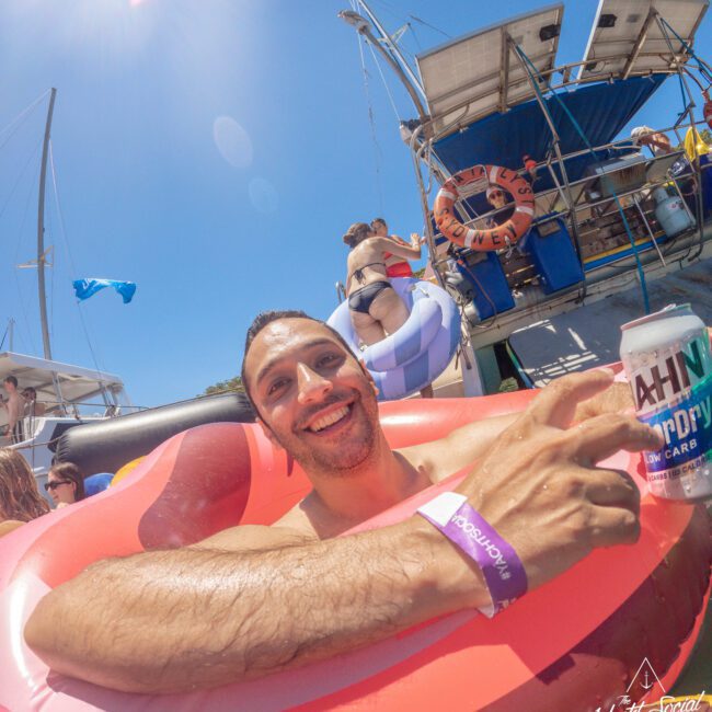 A man smiles while relaxing in a red inflatable float on the water, holding a can of drink. Other people are seen on a boat and in floats nearby under a clear, sunny sky.
