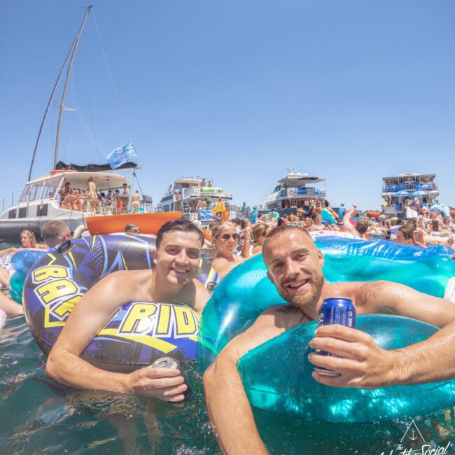 Two men in colorful pool floats hold drinks and smile at the camera while floating in the water, surrounded by other people and anchored yachts under a clear blue sky at a lively boat party.