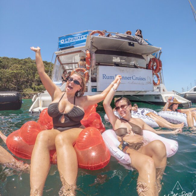 Two women in swimsuits laugh and relax on inflatable floats in the water near a Rum Runner Cruises boat, with other people enjoying the sunny day and clear blue sky in the background.
