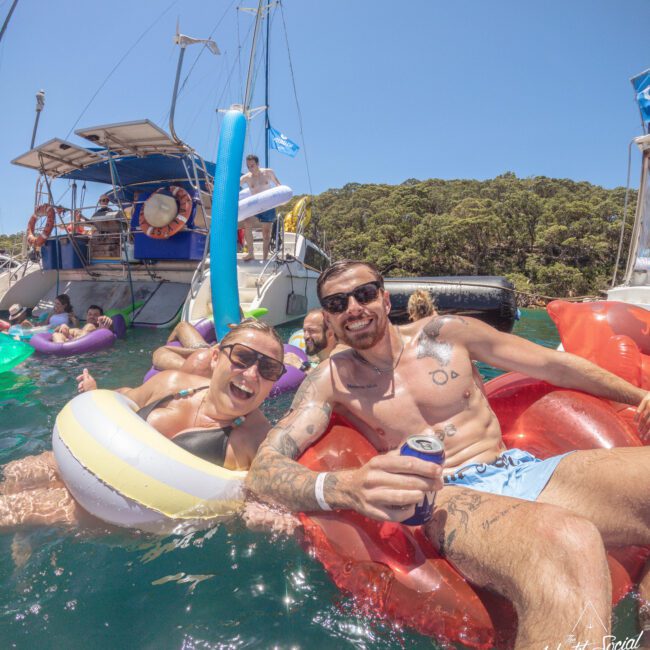 Two people smiling and relaxing on inflatable floats in the water near a boat, surrounded by others enjoying a sunny day. Both are wearing swimsuits, sunglasses, and holding drinks, with trees visible in the background.