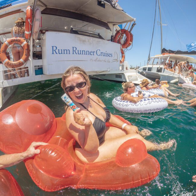 A woman in sunglasses and a black bikini smiles and poses on a red inflatable float in the water near a boat with a "Rum Runner Cruises" banner. Other people relax on floats and enjoy the sunny day by the yacht.