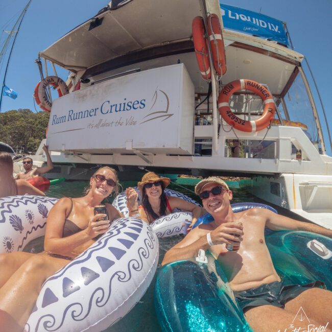 Three smiling people relax on floaties in the water, holding drinks, near a boat labeled "Rum Runner Cruises" on a sunny day. The scene looks lively and cheerful, with blue skies and green trees in the background.