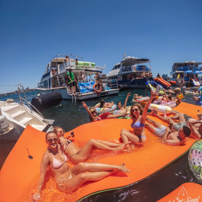 A group of people in swimsuits relax and pose on a large orange float in the water, surrounded by boats and others enjoying a sunny party. The scene is lively and festive under a clear blue sky.