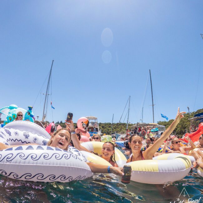 A group of people float on inflatable pool rings in the sea, smiling and raising drinks, with sailboats and a sunny sky in the background. The scene is lively and festive.