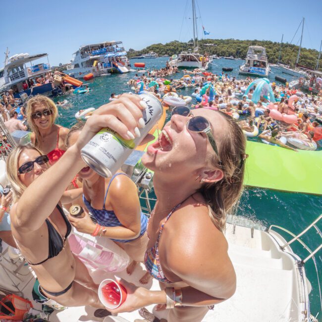 A woman in sunglasses pours a drink from a can into her mouth on a boat, surrounded by friends in swimwear. Behind them, a crowded pool party with floats and boats is underway on a sunny day.