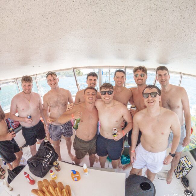 A group of nine smiling men, all shirtless and wearing swim trunks, stand together on a boat holding drinks. There are hot dog buns and snacks on a table in front of them. The ocean is visible through the windows behind them.