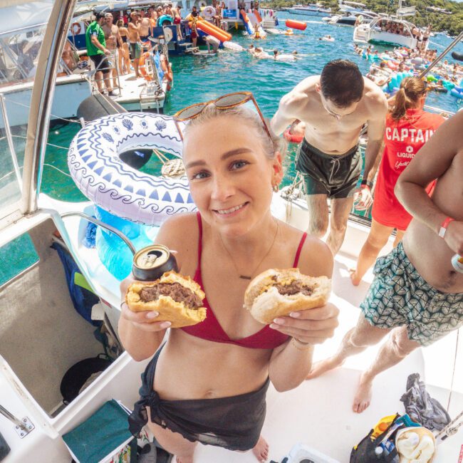 A woman in a red bikini top and black shorts holds two burgers and a can on a boat, smiling at the camera. Other people enjoy the sunny party atmosphere on boats and in the water behind her.