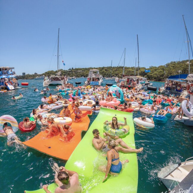 A lively scene of people relaxing and swimming on colorful floats and mats, surrounded by boats and yachts on a sunny, blue-water bay, with greenery in the background. The event looks festive and crowded.