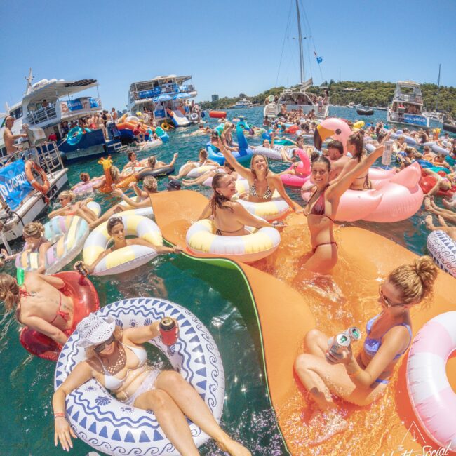 A group of people in swimsuits relax on colorful pool floats and inflatables in clear blue water, surrounded by boats and others enjoying a lively summer party under the sun.
