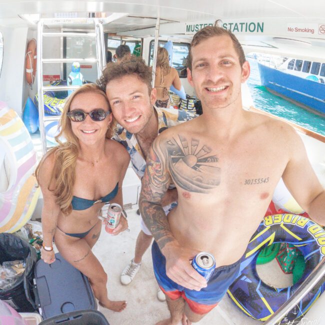 Three smiling adults in swimwear stand on a boat with drinks in hand. Behind them are pool floats, more people, and a blue boat in the water. Sunlight fills the scene, creating a lively, tropical atmosphere.