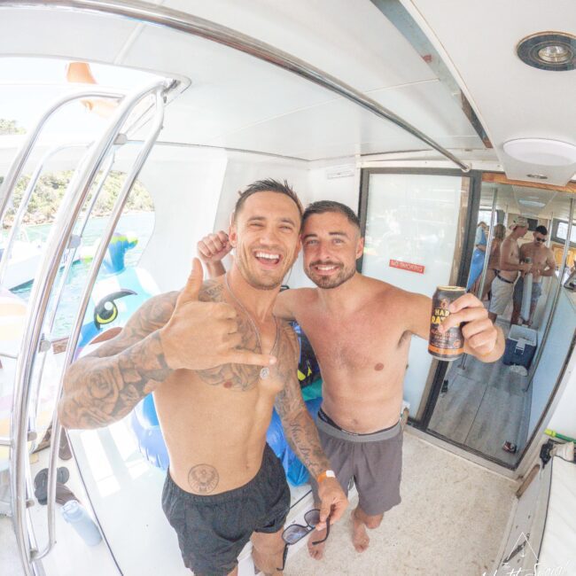 Two men smiling and posing shirtless on a boat, one holding a drink and the other giving a shaka hand sign. A pool float and other people are visible in the background on a sunny day.