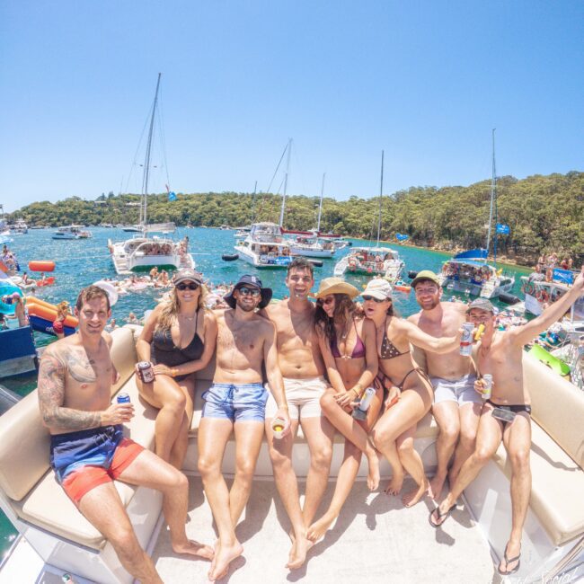 A group of seven young adults in swimsuits sit closely together, smiling and posing on the back of a boat. In the background, boats float on a blue lake surrounded by green trees on a sunny day.