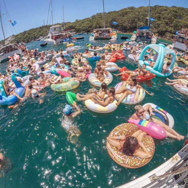 A large group of people relax on colorful inflatable pool floats in the water near several anchored boats, enjoying a sunny day. The scene is lively and festive, with trees visible in the background.