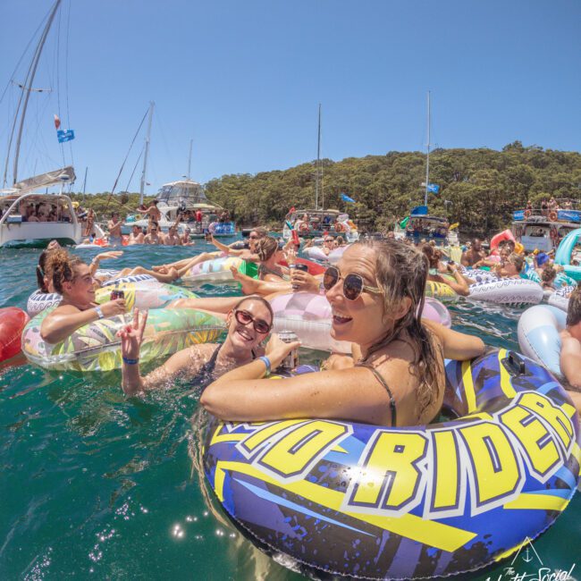 A group of people relax and smile in inflatable tubes on the water near anchored boats, enjoying a sunny day. The foreground shows three women in floats, laughing and posing for the camera.