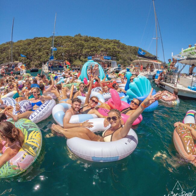 A large group of people relax and smile on colorful inflatable pool floats in the water, surrounded by boats, on a sunny day with clear blue skies and trees in the background.