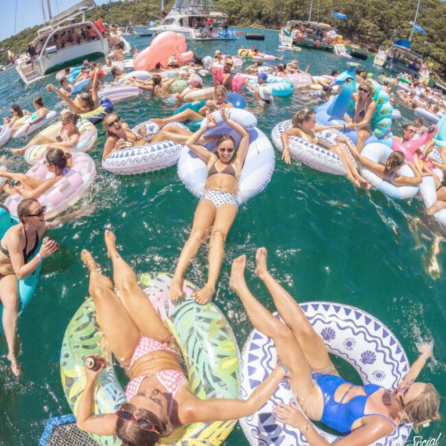 A large group of people relax on colorful inflatable floats in the water near boats, enjoying a sunny day. Many are wearing swimsuits, laughing, and socializing, creating a lively, festive summer atmosphere.