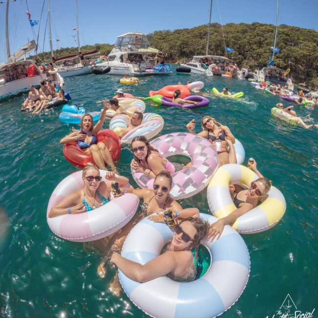 A group of people relax on colorful inflatable rings in clear blue water near docked yachts, enjoying drinks and sunny weather, surrounded by trees and more floats.