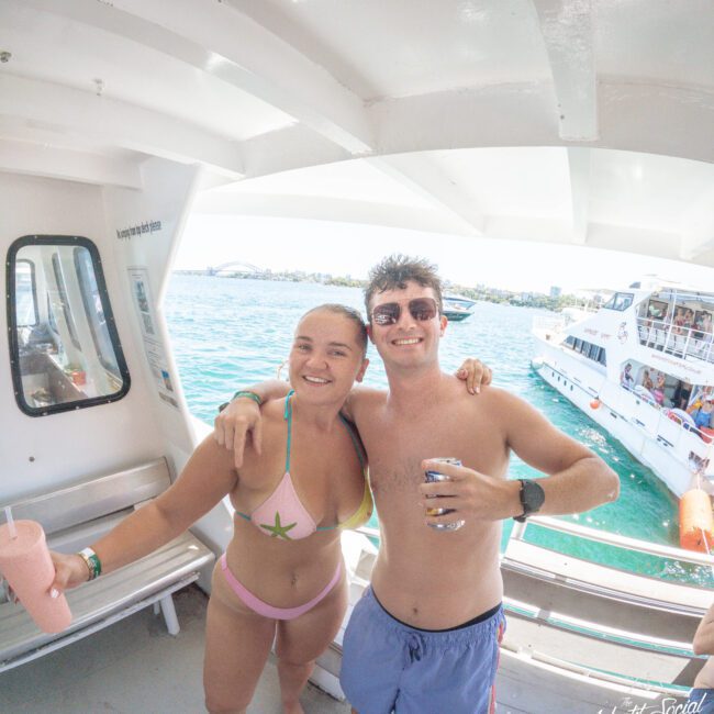 Two people in swimwear smile and pose for a photo on a boat, holding drinks. Bright turquoise water and another boat are visible in the background under a sunny sky.