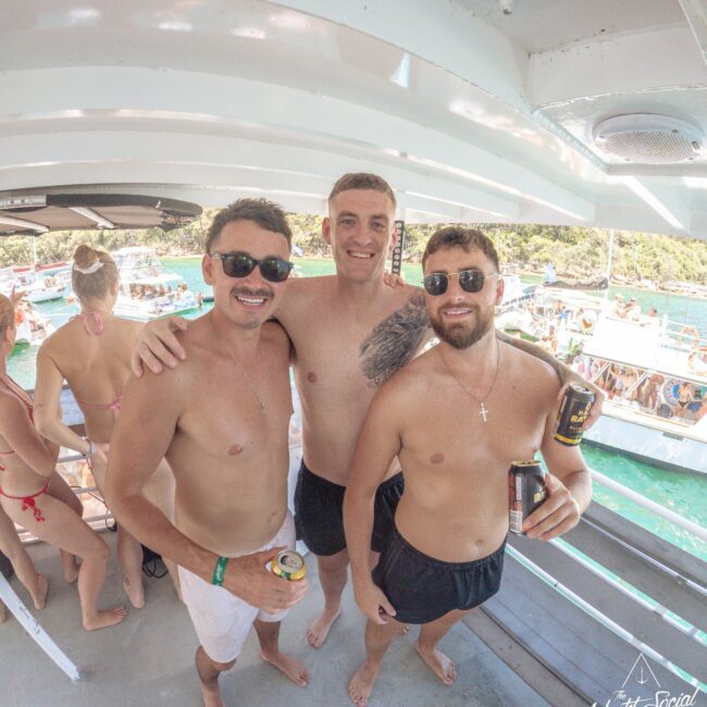 Three smiling men in swimsuits and sunglasses stand on a boat deck, holding drinks. Other people are socializing and swimming in the turquoise water in the background, with trees and more boats visible.