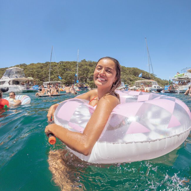 A smiling woman in a pink and white inflatable ring floats in clear blue water near several boats, with people relaxing and trees in the background under a sunny sky.