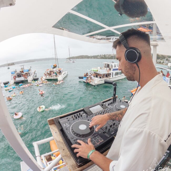A DJ wearing headphones mixes music on a boat deck overlooking a lively scene of people swimming and relaxing on floats near other boats in a bay. The water is blue-green, and the atmosphere is festive and relaxed.