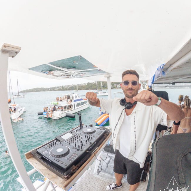 A DJ wearing sunglasses and headphones smiles and poses with raised fists on a boat deck, with a DJ controller in front of him and people on boats in the water in the background.