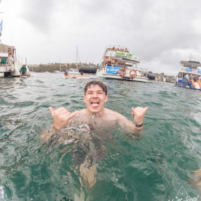 A young man smiles and makes shaka hand signs while swimming in the sea, with several anchored boats and people in the background under a cloudy sky.
