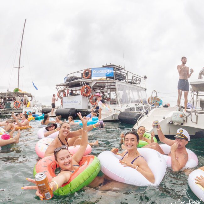 A group of people in colorful floaties smile and hold drinks while partying in the water near several docked boats. The atmosphere is festive, with more people relaxing and celebrating on the boats in the background.