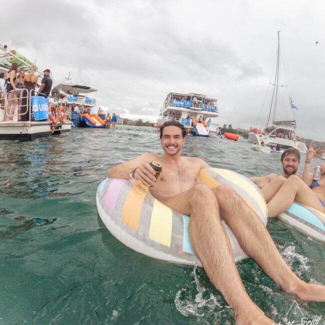 A man smiles while lounging on a colorful inflatable ring, holding a drink in the water near boats. Other people are visible in the background, enjoying a lively, overcast day on the water.