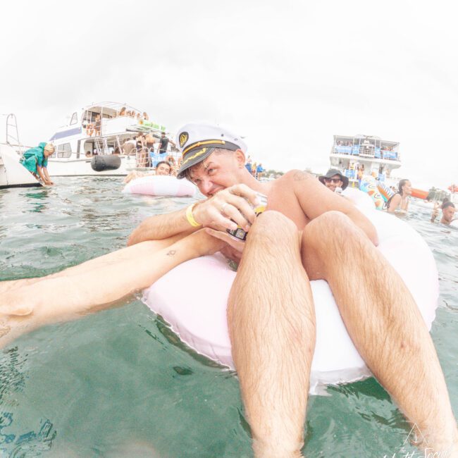 A man wearing a captain's hat lounges on a pink float in the water, holding a drink and playfully biting someone's leg. Boats and people are visible in the background during a lively gathering.