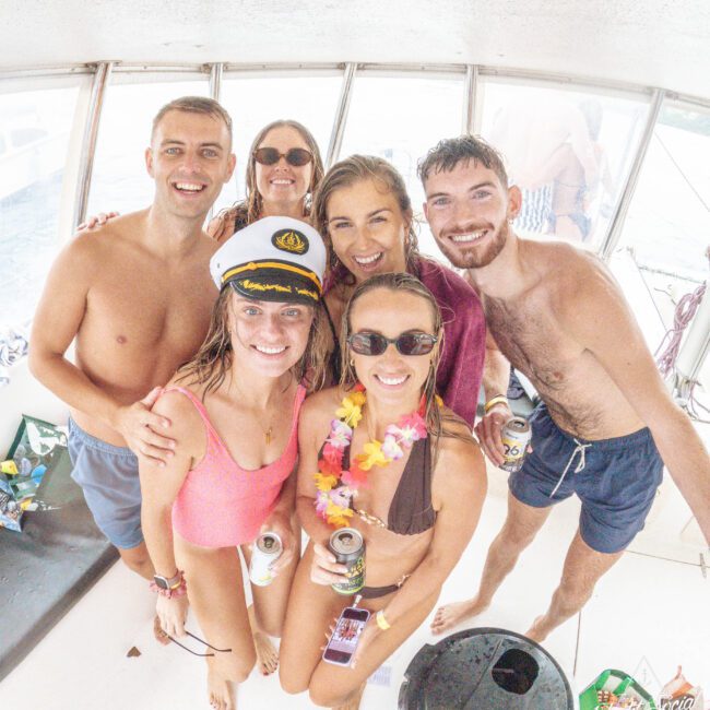 Six smiling young adults pose together on a boat, wearing swimsuits and holding drinks. One woman wears a captain's hat, another a lei. It's bright and lively, with water visible in the background.