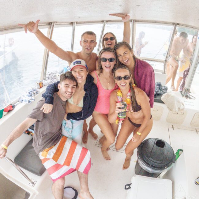 A group of seven smiling young adults in swimsuits pose for a photo together on a boat, with tropical accessories and towels, appearing joyful and relaxed as they enjoy their day on the water.