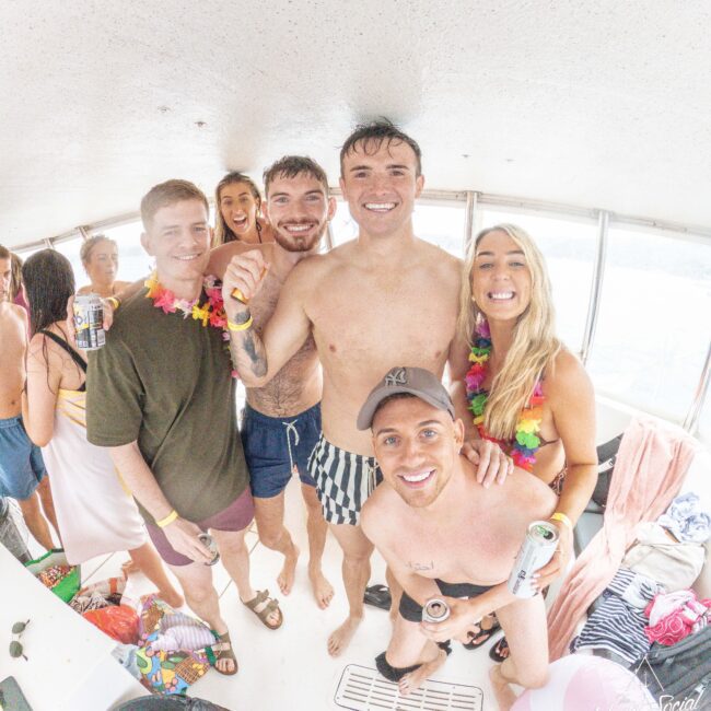 A group of smiling young adults in swimsuits and leis pose together on a boat. Some hold drinks, and there are towels and beach items around them. Sunlight streams through the boat windows, creating a cheerful, festive atmosphere.