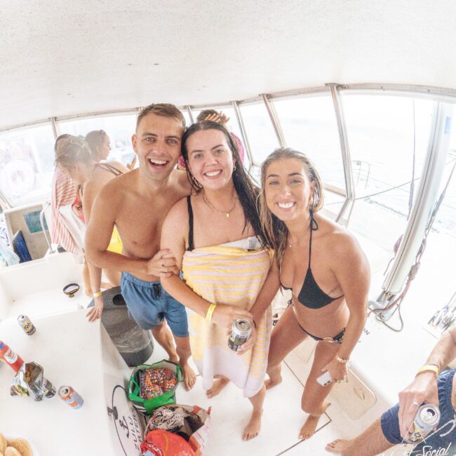 Three smiling people in swimsuits pose together on a boat, one wrapped in a towel, holding drinks. The background shows other people, snacks, and cans, creating a lively, fun atmosphere.