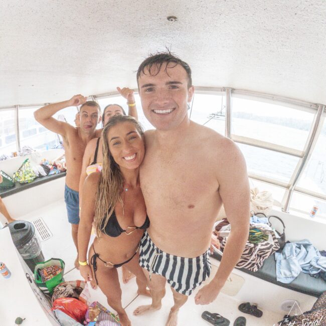 Four smiling young adults in swimsuits pose together on a boat, with a view of water outside the windows. Towels, bags, and snacks are scattered around, and the atmosphere is relaxed and cheerful.