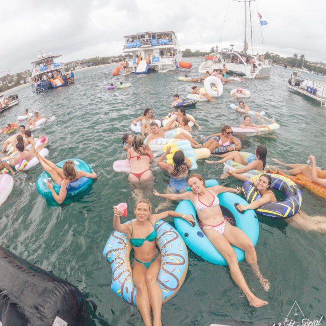 A group of people float on colorful inflatables in a lively party scene on the water, with boats anchored nearby. Most wear swimsuits and appear to be relaxing or celebrating under a cloudy sky.