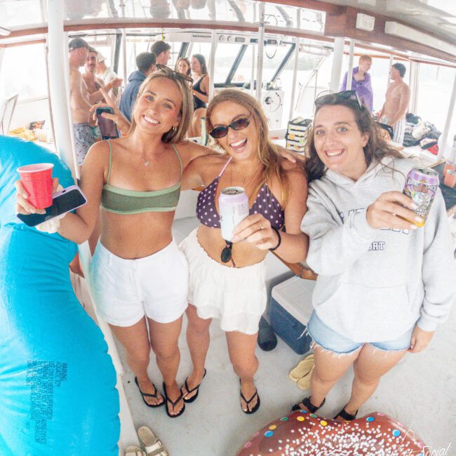 Three women smiling and holding drinks on a boat, surrounded by pool floats and other people enjoying a lively party atmosphere.