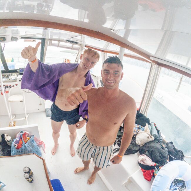 Two smiling men in swim trunks stand on a boat, giving thumbs up. The man on the left has light hair and wears a purple towel, while the man on the right has dark hair and striped shorts. The boat interior is bright and beach gear is visible.