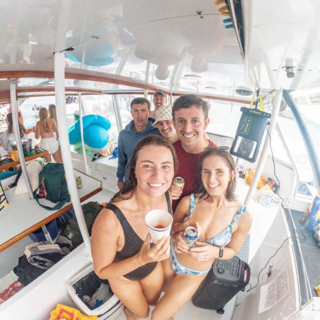 A group of five smiling young adults pose together on a boat, holding drinks. The boat is lively with other people, pool floats, and various gear in the background, creating a cheerful, social atmosphere.