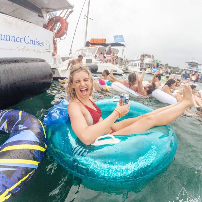 A woman in a red swimsuit lounges on an inflatable tube in the water, laughing and holding a drink, surrounded by other people and boats at a lively party.